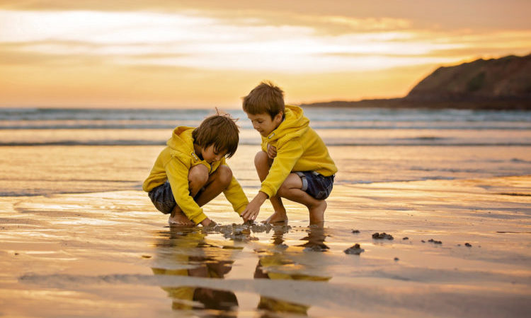 Two beautiful children, boy brothers, playing on the beach with sand and running in the water on sunset, Devon, England